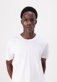 Tricou alb din bumbac, cu guler rotund și mâneci scurte, purtat de un model cu părul scurt și texturat, pe un fundal neutru.