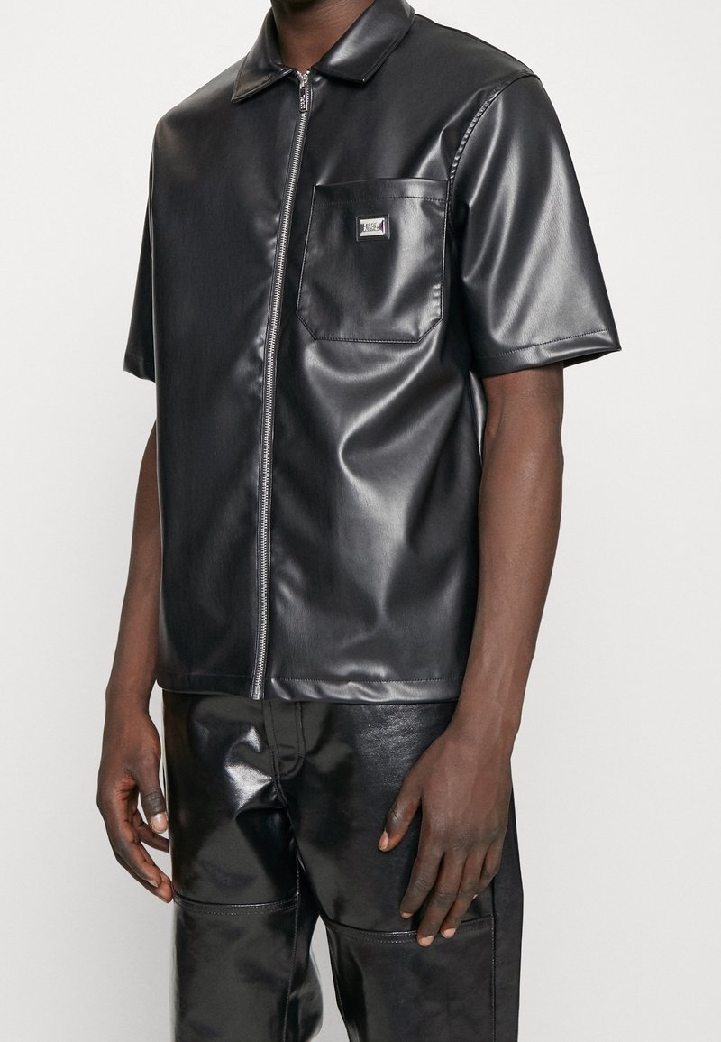 Zwart shirt met korte mouwen van imitatieleer, voorzien van een ritssluiting, puntkraag en borstzakje. Gladde textuur met een glanzende afwerking.
