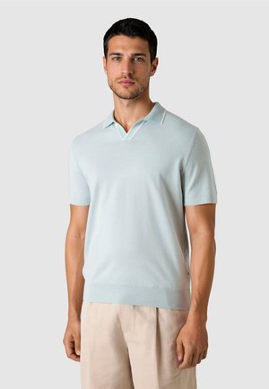 Junger Mann trägt ein hellblaues Polohemd mit kurzen Ärmeln und beige Hose, steht vor einem schlichten grauen Hintergrund.