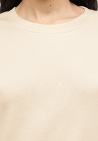 Haut en tissu beige côtelé avec un col rond. La texture est lisse et douce, présentant de fines lignes verticales. Pas de motifs ou accents distincts.