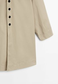 Chemise beige à manches longues avec une fermeture à boutons à l'avant, dotée de boutons noirs, d'une texture lisse et d'une coupe décontractée.