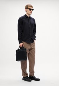 Svart långärmad skjorta med texturerat mönster, beige byxor, svarta skor och en svart läderväska, accessoriserad med mörka solglasögon.