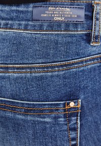 Blå denimbukser har oransje sømmer, en baklomme og en etikett med tekst som angir merke og stil, plassert ved midjen.
