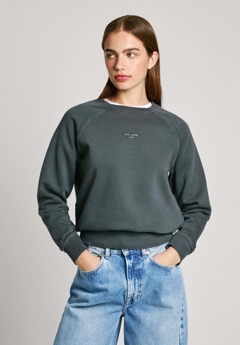 Mörkgrå sweatshirt med ribbad mudd och nederkant, utrustad med en liten vit logotyp på bröstet, tillsammans med ljusblå jeans.