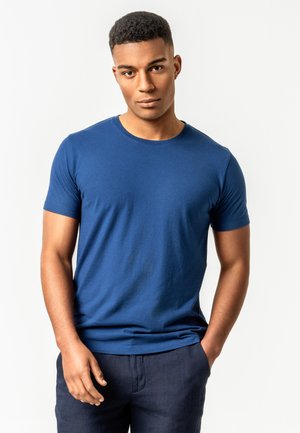 Junger Mann mit kurzen lockigen Haaren, der ein blaues T-Shirt mit Rundhalsausschnitt und dunkle Hosen trägt, steht mit einer Hand in der Tasche vor einem einfarbigen Hintergrund.