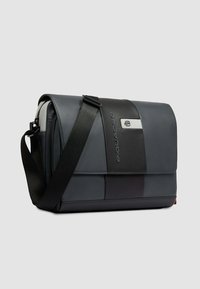 Grijze en zwarte Piquadro leren schoudertas met verstelbare zwarte schouderriem en klepafsluiting op lichtgrijze achtergrond.