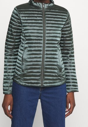 Person trägt eine glänzende grüne, horizontal gesteppten Reißverschlussjacke mit Taschen und blaue Jeans, dargestellt von Hals bis Hüften.