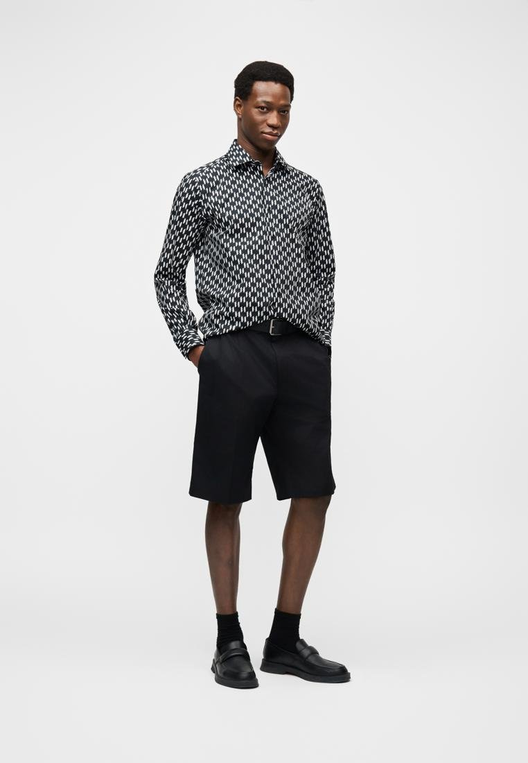 Homme portant une chemise à manches longues noire et blanche à motifs, un short noir ajusté, une ceinture noire, des chaussettes noires et des mocassins noirs, debout les mains dans les poches.
