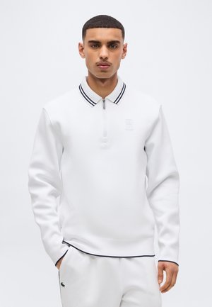 Homme aux cheveux courts portant un polo à manches longues blanc avec des bordures noires et un pantalon blanc, debout devant un fond clair uni.