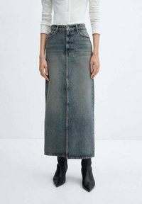 Denim maxi sukně ve vybledlé modré barvě, s vysokým pasem, rovnou siluetou, předními knoflíky a minimalistickým designem. Kombinováno s černými špičatými kozačkami.