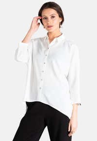 Camisa blanca de botones con cuello, mangas largas y dobladillo asimétrico. Cuenta con botones brillantes y una tela suave y ligera.