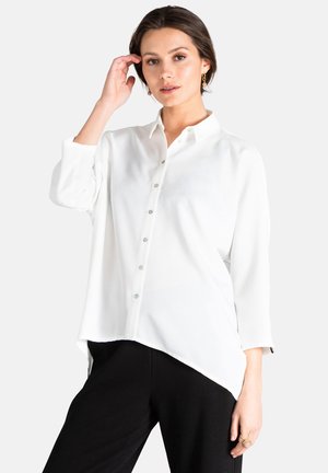 Chemise blanche à boutons avec col, manches longues et ourlet asymétrique. Dotée de boutons brillants et d'un tissu lisse et léger.