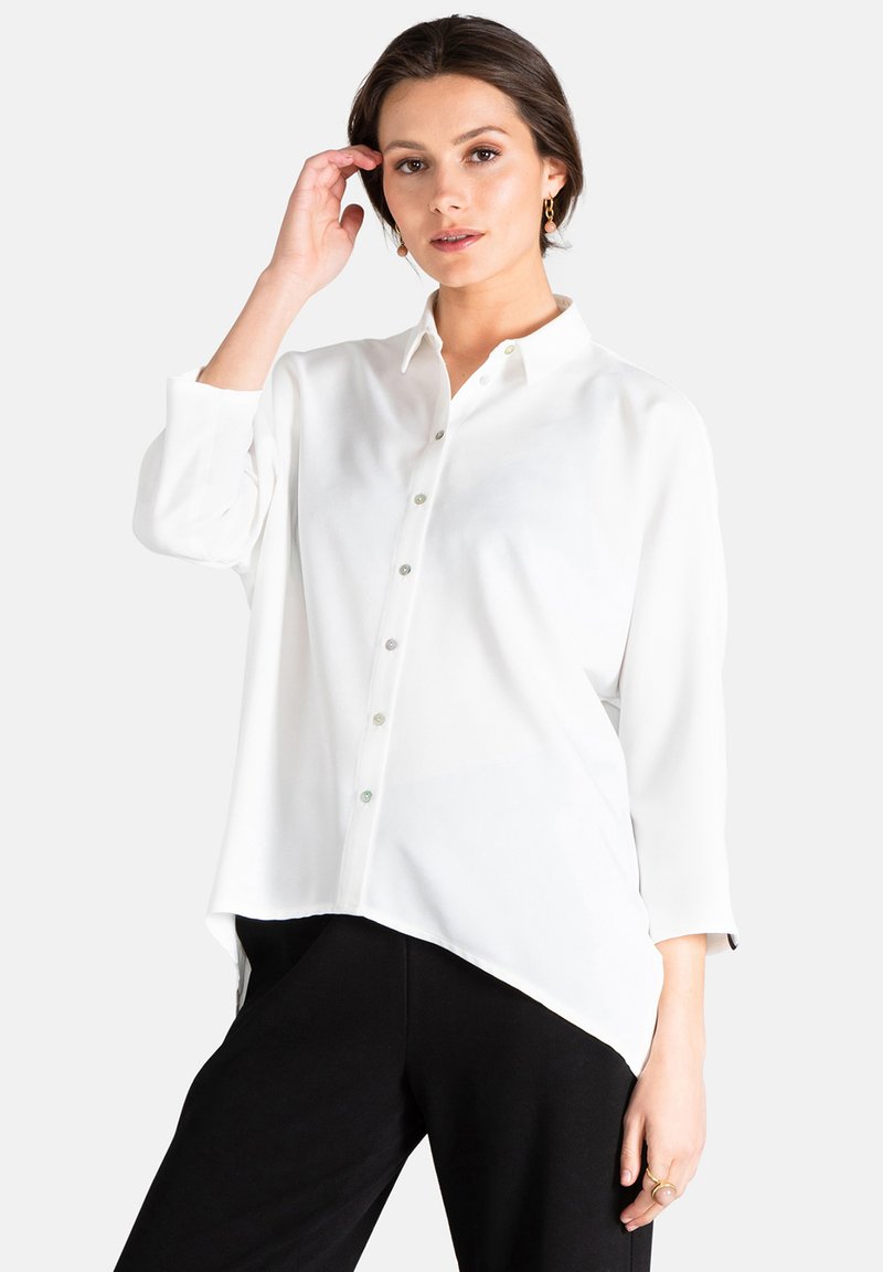 Camisa blanca de botones con cuello, mangas largas y dobladillo asimétrico. Cuenta con botones brillantes y una tela suave y ligera.