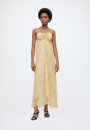 Γυναίκα με φυσικά μαλλιά, που φοράει ένα ανοιχτό κίτρινο φόρεμα μακρύ με λουλούδια και λεπτές τιράντες, καθώς και μαύρα πέδιλα με τακούνι, στέκεται μπροστά σε ένα απλό φόντο.