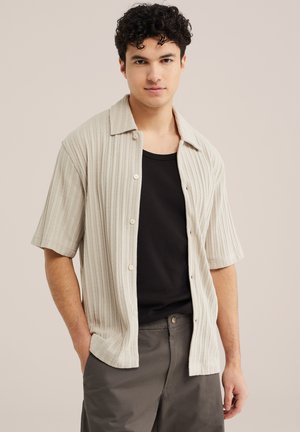 Mladý muž s kučeravými vlasmi, oblečený v béžovej rebrovanej košeli s krátkym rukávom na gombíky, cez čierne tričko a sivé nohavice, na jednoduchom pozadí.