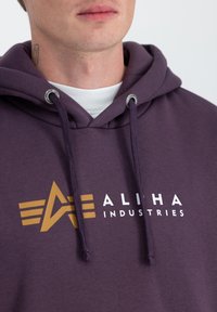 Fialová mikina s kapucňou a šnúrkami, ktorá má zlaté logo "ALIHA INDUSTRIES" a grafický dizajn s horizontálnymi pruhmi na hrudi.