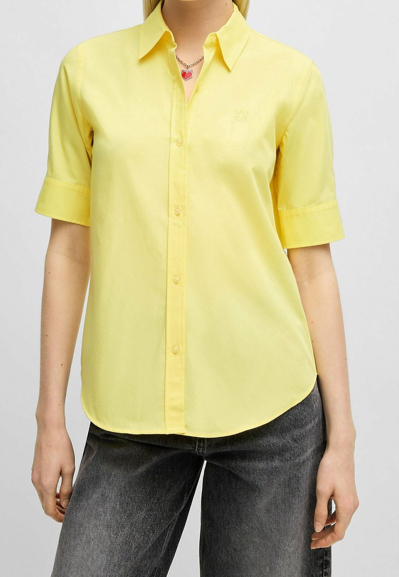 Femme portant une chemise jaune à manches courtes boutonnée, un jean noir et un collier avec un pendentif rouge en forme de cœur.