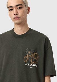 Ciemnozielony bawełniany t-shirt z haftowanym wzorem węża w kolorze złotym i czarnym, z napisem "ALLSAINTS LONDON" na dole.