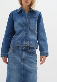 Denim jakna i suknja u srednje plavoj boji. Jakna ima kopčanje na dugmad, džep na prsima i široke rukave. Suknja ima klasičan kroj s prednjim šavom.