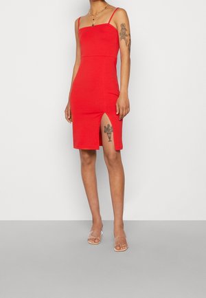 Femme portant une robe rouge sans manches, longueur genoux, avec des bretelles fines et une fente sur la cuisse, montrant des tatouages floraux sur le bras et la jambe, avec des talons à brides transparentes.