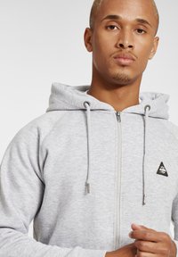 Sweat à capuche gris avec fermeture éclair sur le devant, cordons de serrage gris, texture du tissu et logo triangulaire noir sur la zone gauche de la poitrine.