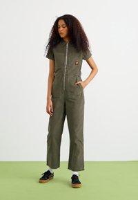 Tuta verde oliva con maniche corte, chiusura zipper, tasche anteriori e vita regolabile. Abbinato a sneakers nere e calzini bianchi.