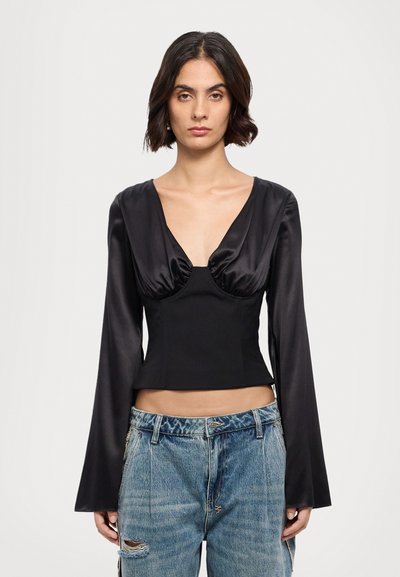 Top lungo in satin nero a maniche lunghe con scollatura a V profonda, caratterizzato da dettagli arricciati sul busto e una silhouette aderente. Abbinato a jeans strappati.