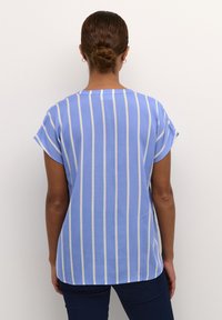 Blouse à manches courtes en bleu clair avec des rayures verticales blanches et crème. Tissu doux avec une coupe ample et un ourlet arrondi.