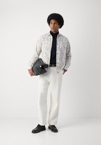 Camisa blanca con estampado de paisley, cuello alto azul marino, pantalones beige, zapatos negros, llevando un bolso negro de gran tamaño. Tejido suave, corte relajado.