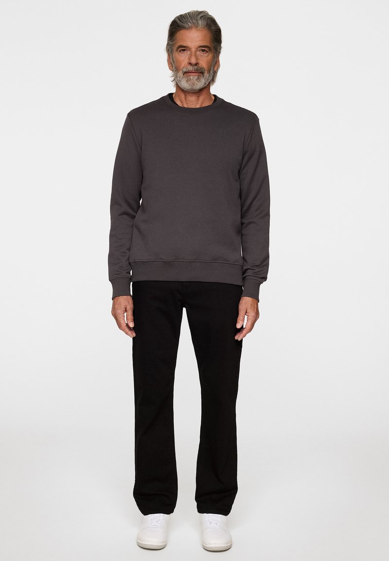 Un sweat-shirt gris avec un col rond, des manches longues et des bords côtelés, associé à un pantalon noir et des baskets blanches, se tenant sur un fond neutre.