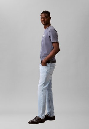 Άνδρας που στέκεται πλάγια, φοράει ξεθωριασμένο μοβ μπλουζάκι, ανοιχτό μπλε τζιν και μαύρα loafers, με το ένα χέρι στην τσέπη, σε απλό φόντο.