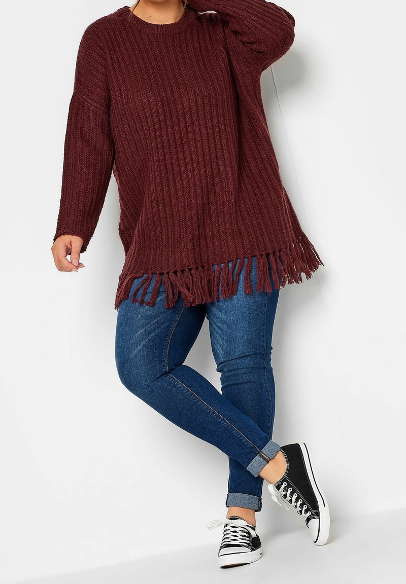 Personne portant un long pull en maille bordeaux avec des franges, un jean skinny bleu retroussé et des baskets basses noires et blanches, posant avec une jambe croisée.