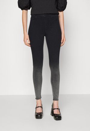 Kobieta w czarnych przechodzących w szare ombre obcisłych jeansach, czarnej bluzce z bufkami i czarnych szpilkach na platformie Mary Jane, stojąca na jednolitym tle.