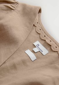 Haut en lin marron avec un détail de col festonné. Les étiquettes incluent "N" et "Premium Collection" en textile blanc. Tissu texturé.