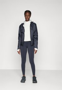 Vrouw in een zwarte sportjack, witte lange mouw top, donkere leggings en zwarte sneakers die tegen een effen grijze achtergrond staat.