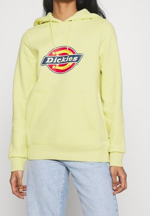 Osoba nosząca jasnofioletową bluzę z kapturem Dickies z przednią kieszenią i kapturem, w zestawieniu z jasnoniebieskimi dżinsami, stojąca na jednolitym tle.