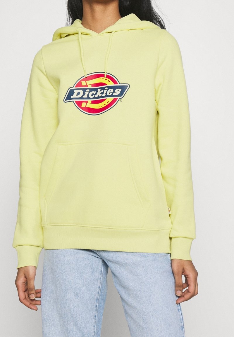 Personne portant un sweat à capuche Dickies jaune clair avec une poche avant et une capuche, associé à un jean bleu clair, debout devant un fond uni.