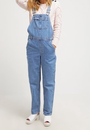 Person iført lyseblå denim- overalls, cremefarvet kabelstrikket sweater og hvide platformssandaler med åben tå, stående med den ene hånd i lommen.