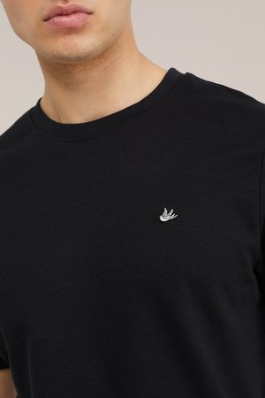 Uomo che indossa una semplice maglietta nera a girocollo con un piccolo logo bianco a forma di uccello sul petto sinistro, su sfondo neutro.
