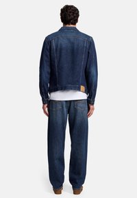 Giacca di jeans blu con un taglio leggermente corto, polsini abbottonati e un'etichetta in pelle beige. Abbinata a jeans blu a taglio largo.