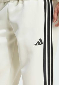 Bijele atletičarske hlače od glatkog materijala s tri crne pruge sa strane i crnim Adidas logom blizu kuka.