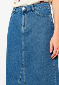 Vidutinio mėlynumo džinsinė midi sijonas su priekine plyšio, klasikinio penkių kišenių dizainu ir metaliniu sagos uždarymu. Audinys turi lygų, tekstūruotą paviršių.