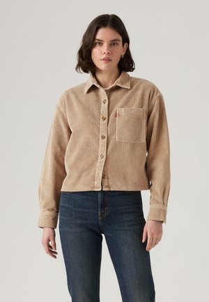 Beige kordfløyelskjorte med knappet front, krage og en enkelt brystlomme, med vertikal ribbet tekstur og lange ermer.
