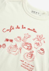 Valkoinen puuvillapaita, jossa on punaisella brodeerattuja kuvastoja kahvilatavarasta, mukaan lukien leivonnaisia, kuppeja ja kukkia. Teksti kuuluu "Café de la nuit."
