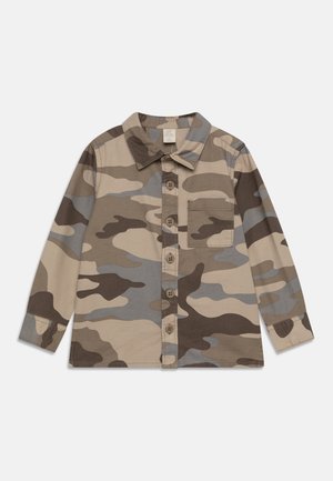 Chemise à manches longues boutonnée avec un motif camouflage beige, marron et gris et une poche poitrine unique, étalée à plat sur un fond blanc.