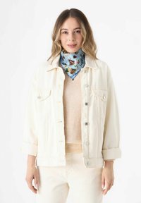 Witte denim jas met opgerolde mouwen, een beige trui eronder en een blauwe sjaal met een patroon van bloemen en vlinderontwerpen.