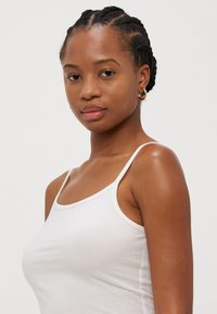 Hvit bomull tanktop med tynne stropper, med en tettsittende design og en jevn tekstur, båret av en modell med flettet hår.