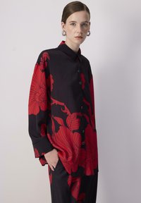 Schwarzes Hemd mit Knöpfen, das große rote Blumendrucke, lange Ärmel und einen entspannten Schnitt aufweist, aus glattem Stoff gefertigt. Sichtbares Taschen-Detail.