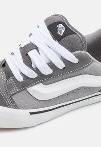 Baskets en daim gris avec lacets blancs, présentant un logo en forme de vague blanche et des accents noirs. Comprend des détails perforés et une semelle en caoutchouc.