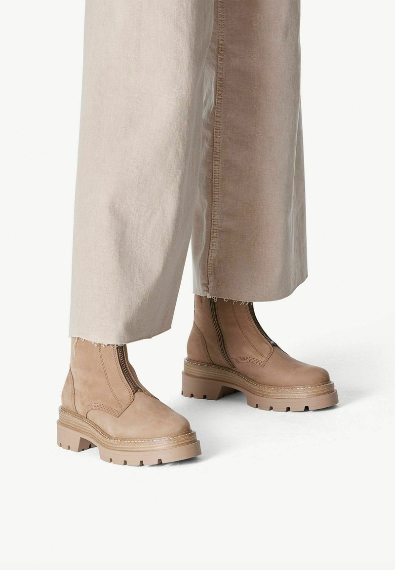 Beige ankle boots with a chunky sole, elastic side panels, and smooth suede texture. Paired with cropped, light-colored pants.